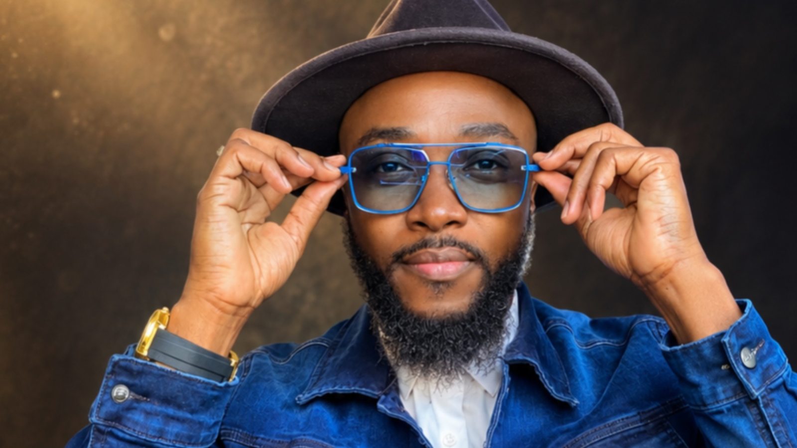 Kevin Downswell in a studio portrait, brown fedora and blue-framed tinted glasses, denim jacket, looking at camera