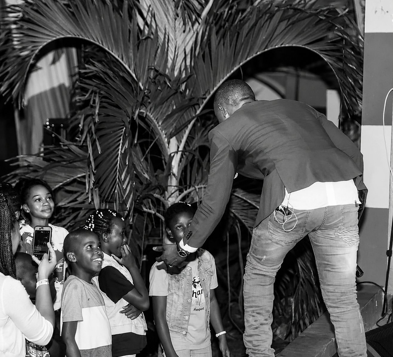 Kevin Downswell leaning down from a stage in black and white to greet a group of children looking up at him, laughing