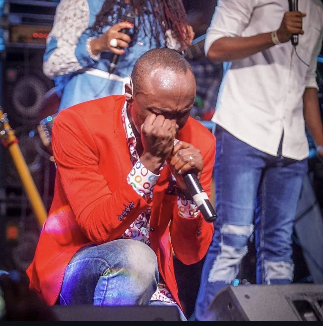 Kevin Downswell crouched on stage in a red jacket, microphone in hand, one hand pressed to his face, overcome in worship